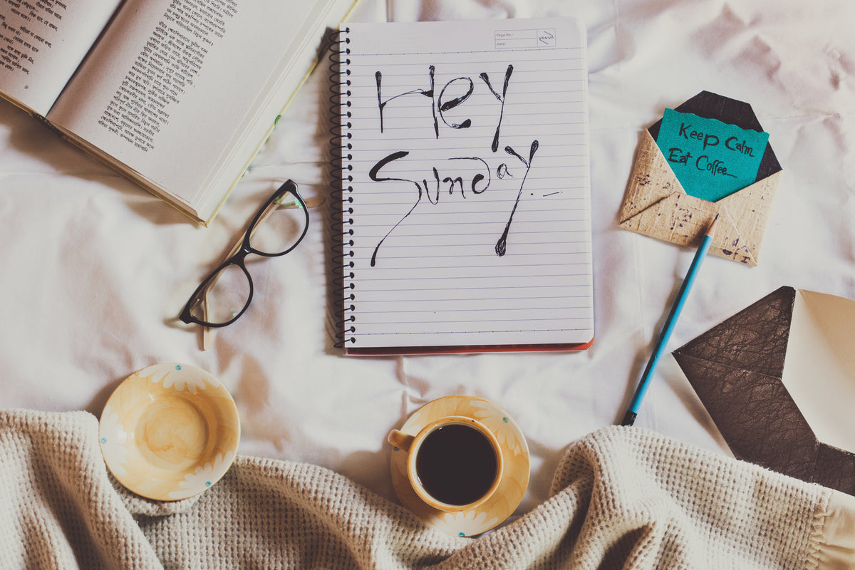 Flatlay of a notebook reading "Hey Sunday" with glasses, coffee, an open book, and an envelope on a bed